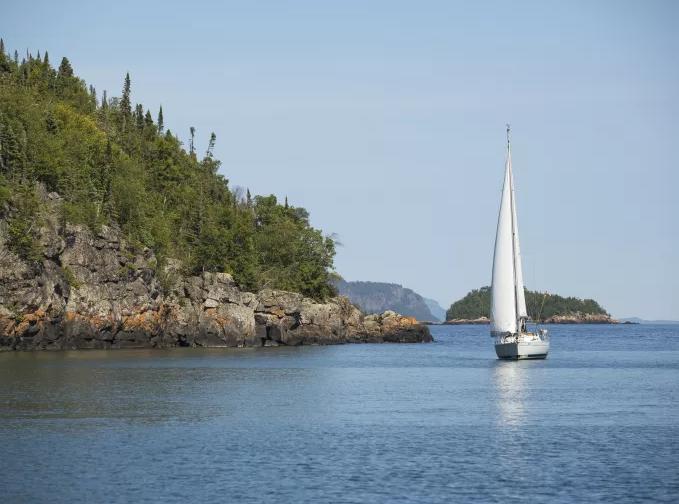 Sailboat gliding near rocky, tree-covered shoreline on a calm lake