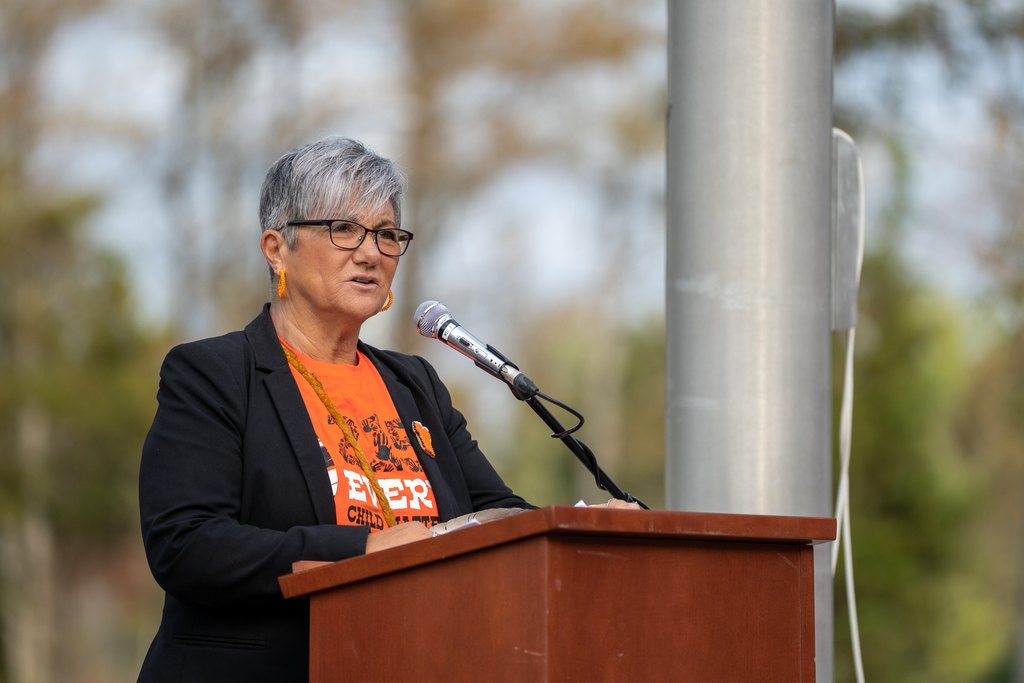 A short-haired person in glasses and a black blazer speaks at a microphone behind a wooden podium outdoors, wearing an orange shirt.