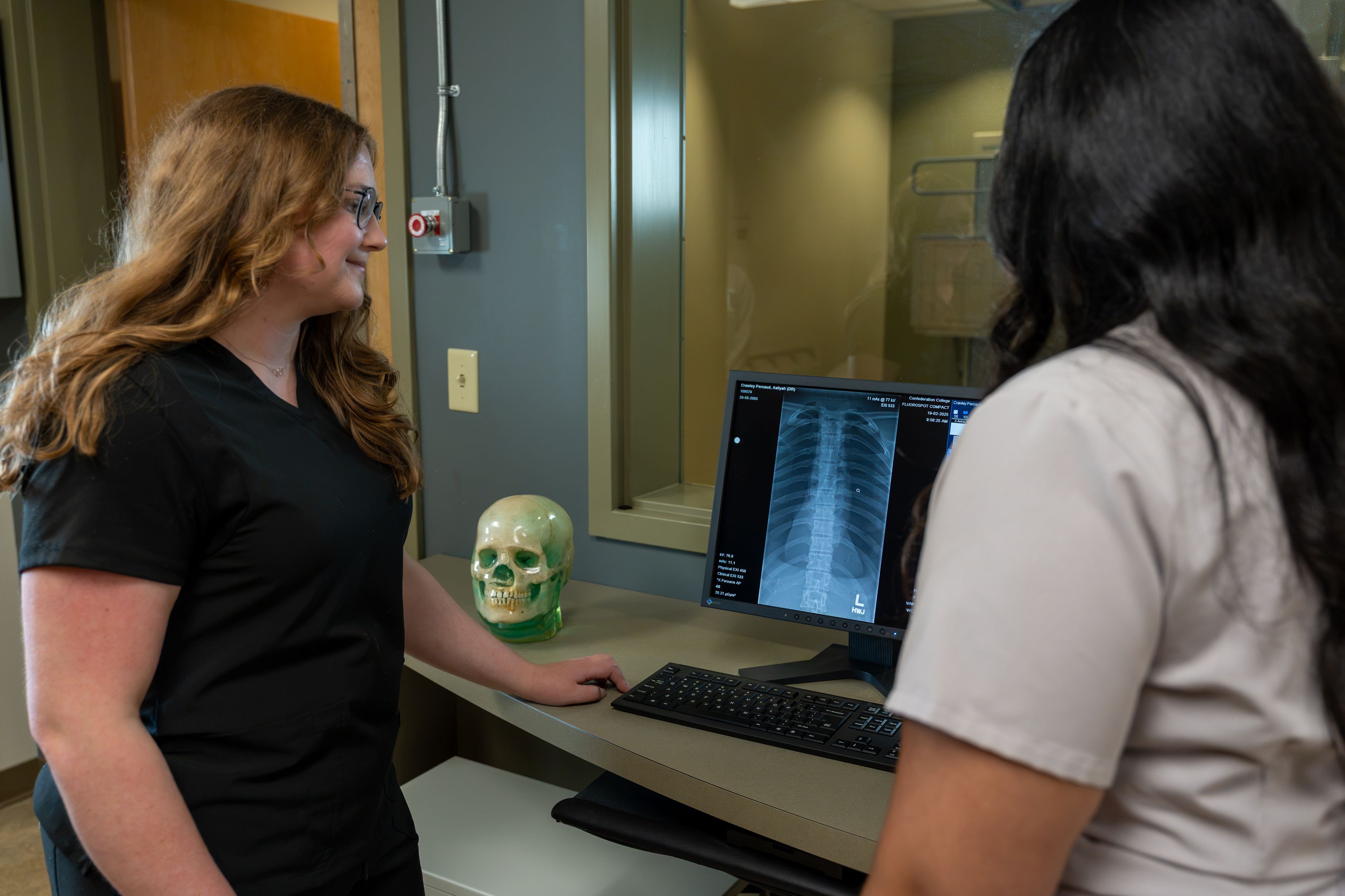Two people stand at a computer displaying a chest X-ray, beside a model skull in a medical imaging room.
