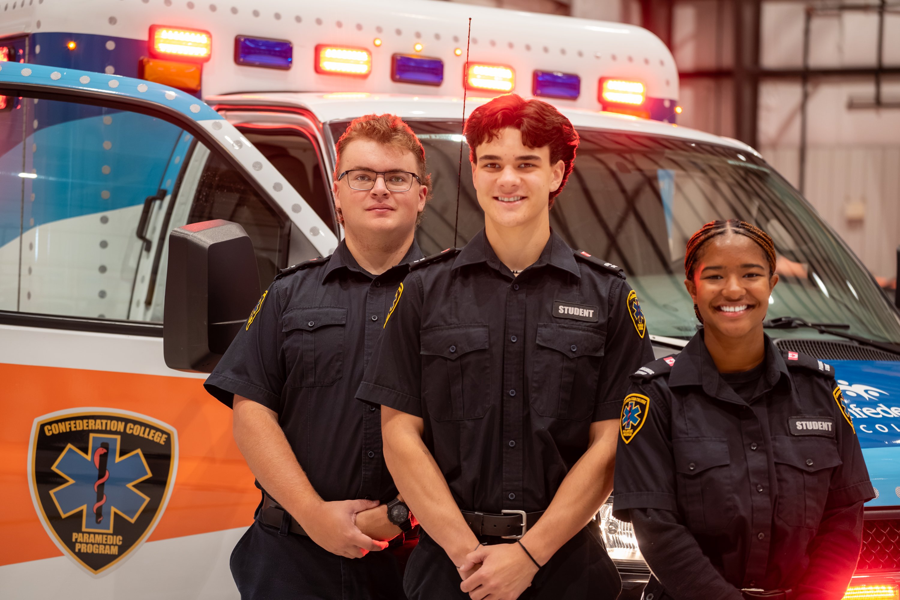 Three paramedic students stand in front of an ambulance with flashing lights and Confederation College Paramedic Program markings.