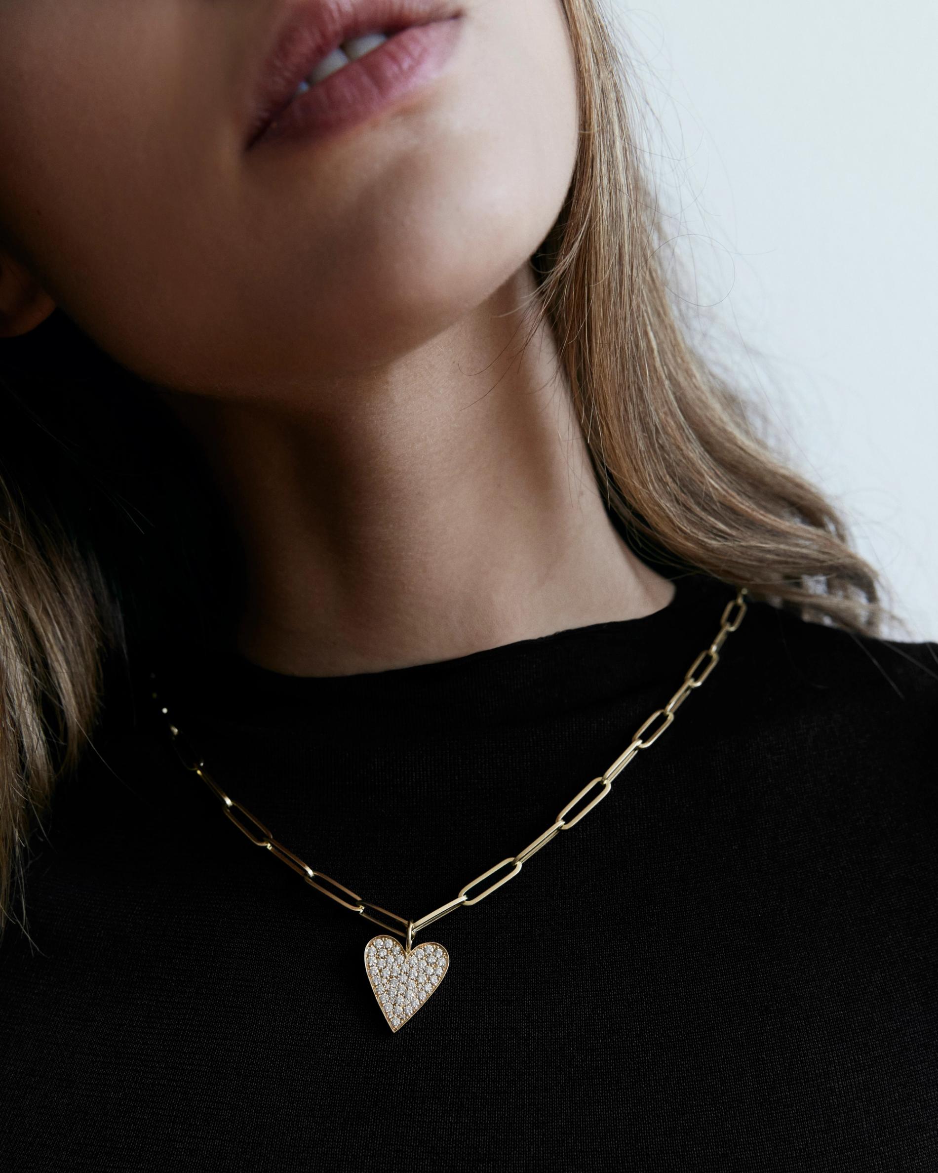 Model wearing large gold chain with diamond heart charm. 