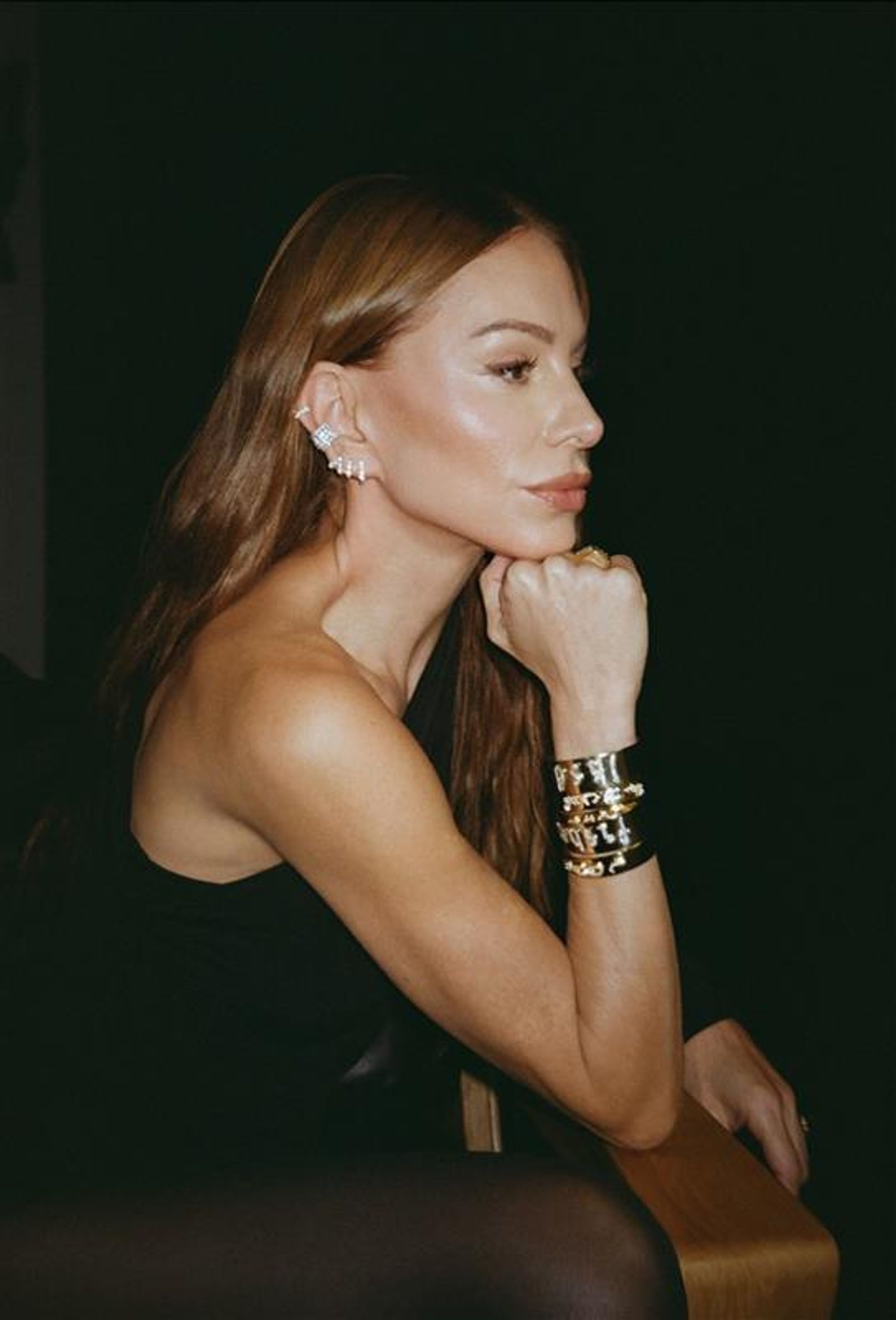 image of jennifer fisher wearing gold bracelets and diamond earrings in a dark room