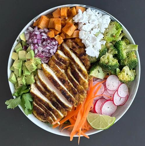Salad bowl of radish, broccoli, vegan feta, sweet potato, red onion, avocado, carrots, cilantro and a lime wedge. Topped with sliced curry chicken.
