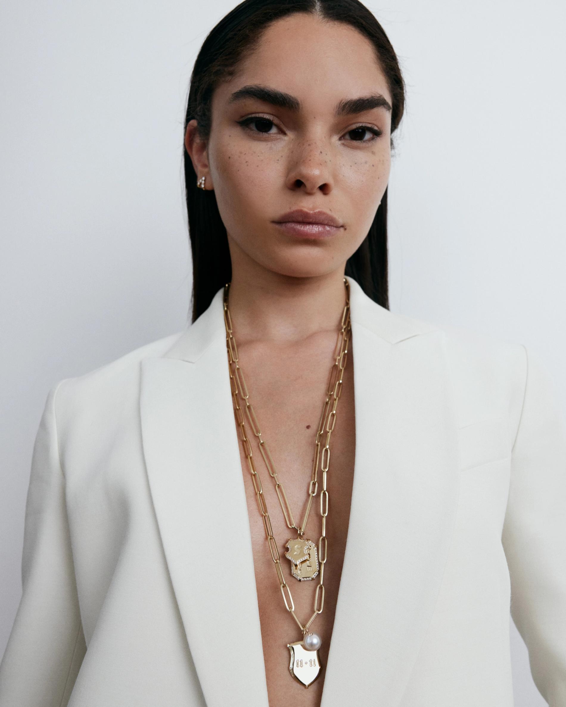 Model with slicked back hair wearing multiple gold chains and assorted charms under white blazer looking directly into camera. 