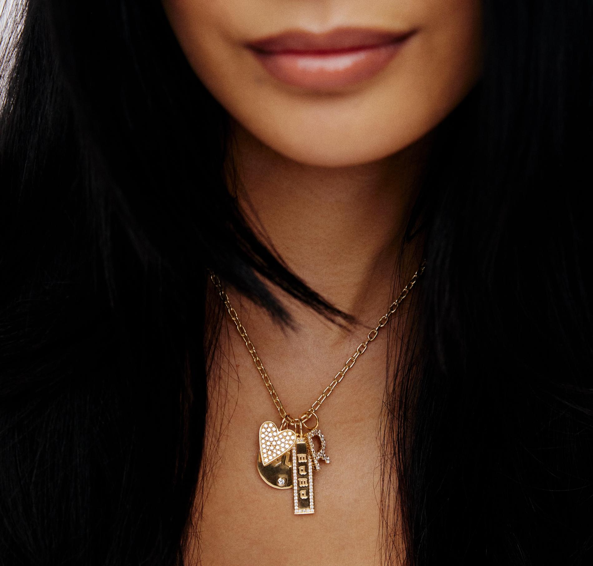model with dark hair wearing a charm necklace on a gold chain with hearts a dog tag and a personalized charm