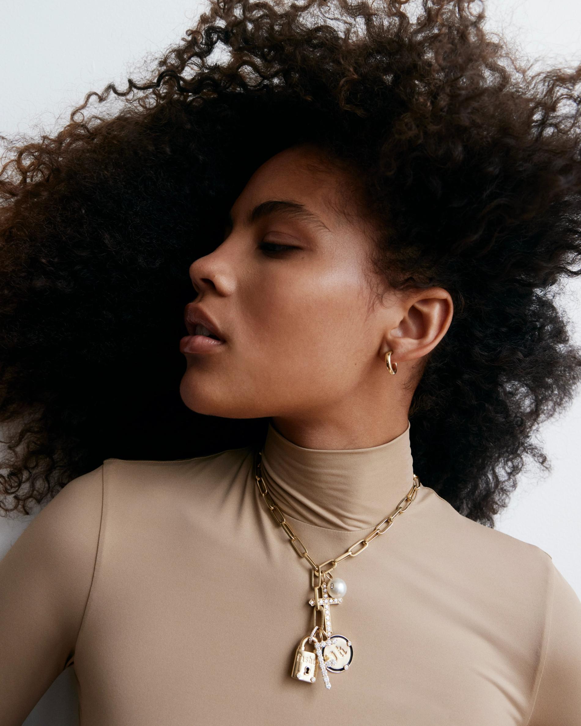 Model wearing beige turtleneck and multiple large gold and diamond charms on a chunky gold necklace 