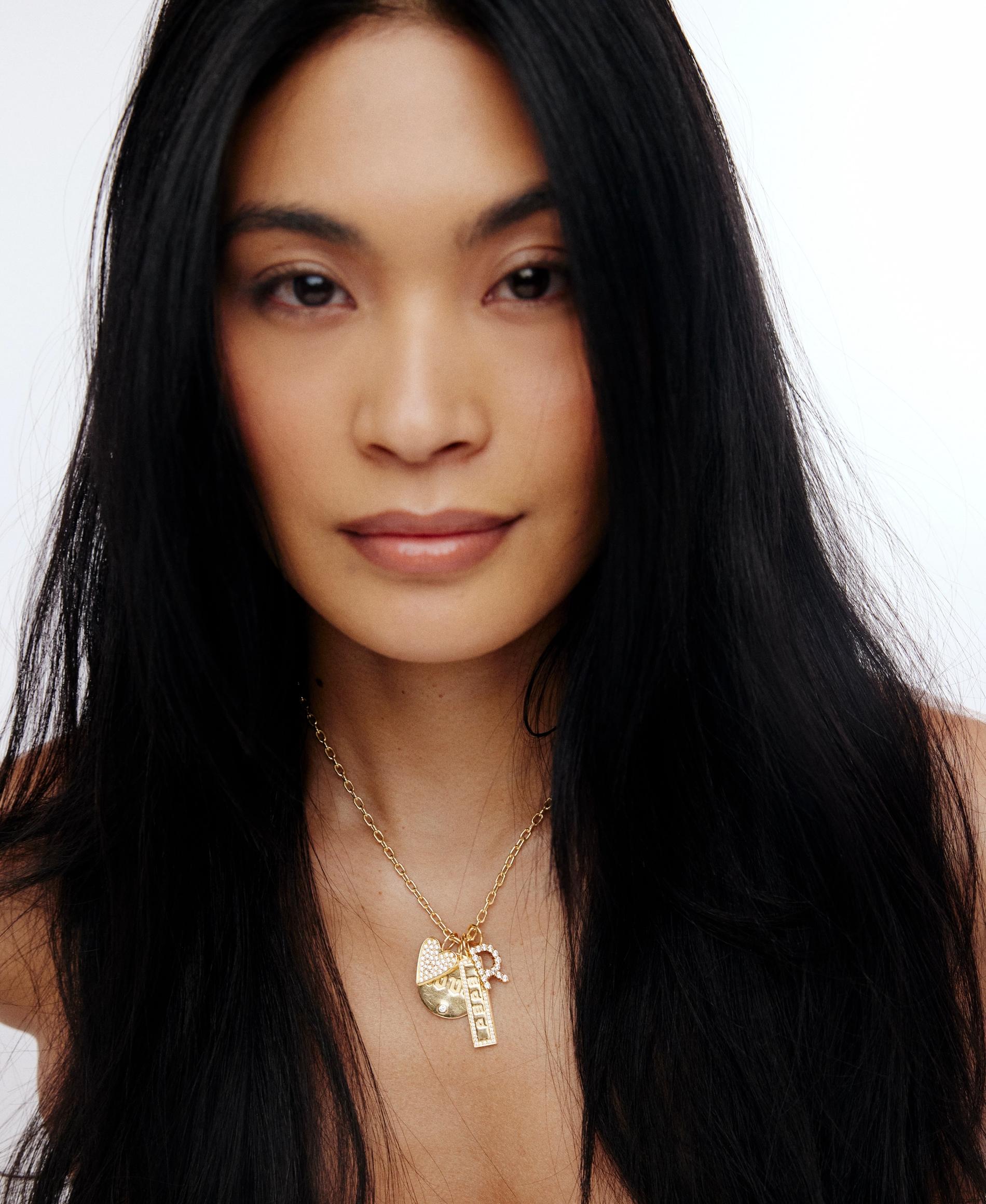 model with dark hair wearing a charm necklace on a gold chain with hearts a dog tag and a personalized charm