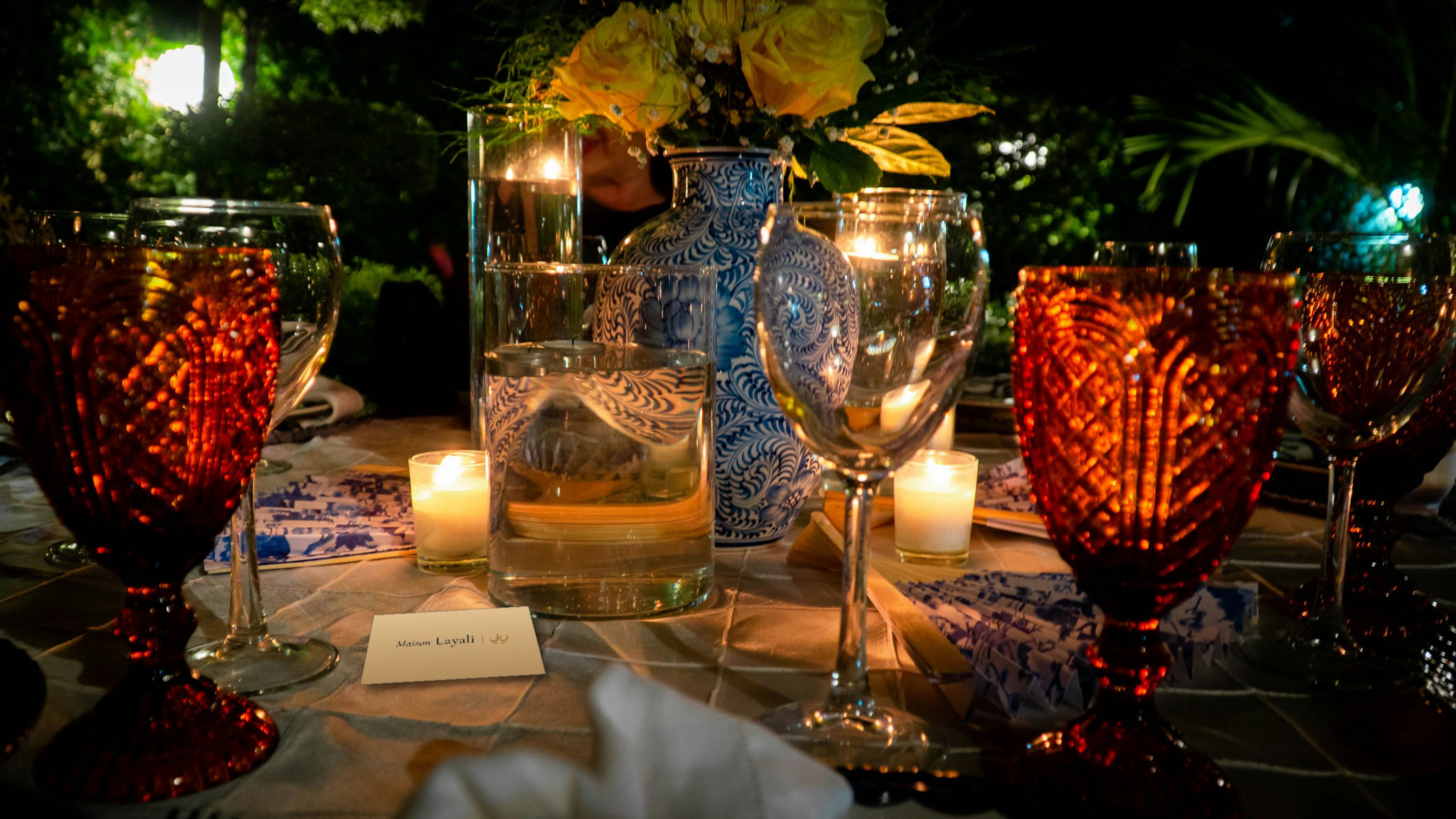 Een gedekte tafel bij avond met amberkleurige glazen, kaarsjes en een blauw beschilderde vaas; op het tafelkleed ligt een kaartje van Maison Layali