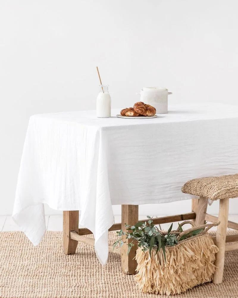 Een tafel met een wit linnen tafelkleed, naast een gevlochten mand met groene takken