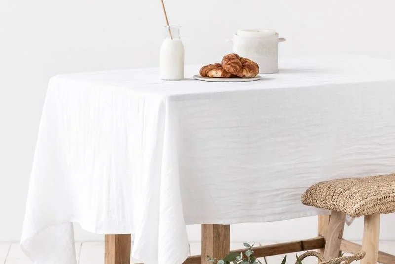 Een tafel met een wit linnen tafelkleed, naast een gevlochten mand met groene takken