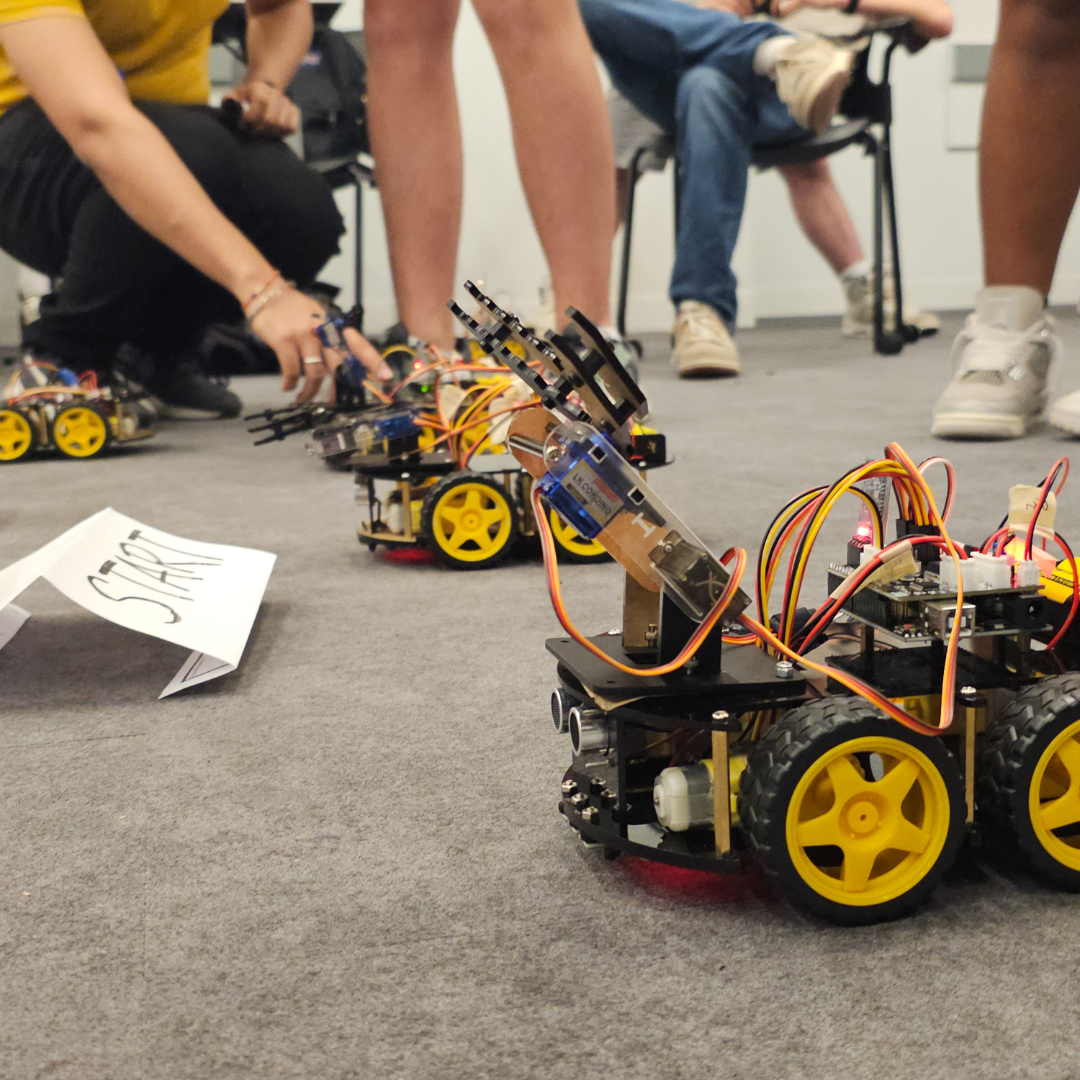 Student-built robotic vehicles with mechanical claws lined up at a starting point for a robotics challenge, with students observing in the background at NYU Tandon’s K12 STEM Center.