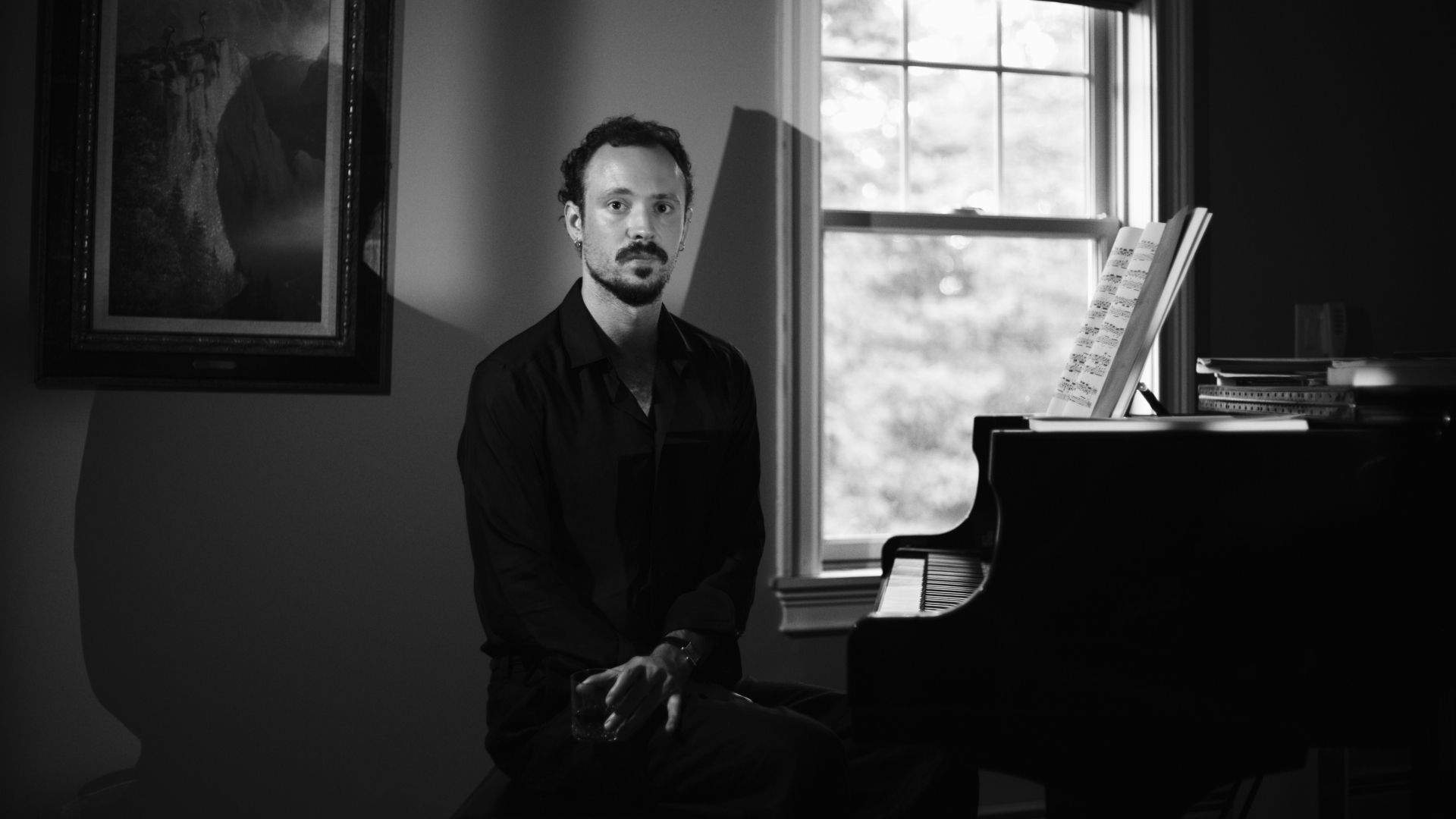 Black-and-white portrait of pianist and composer Eric Christian seated beside a grand piano with sheet music by a window.