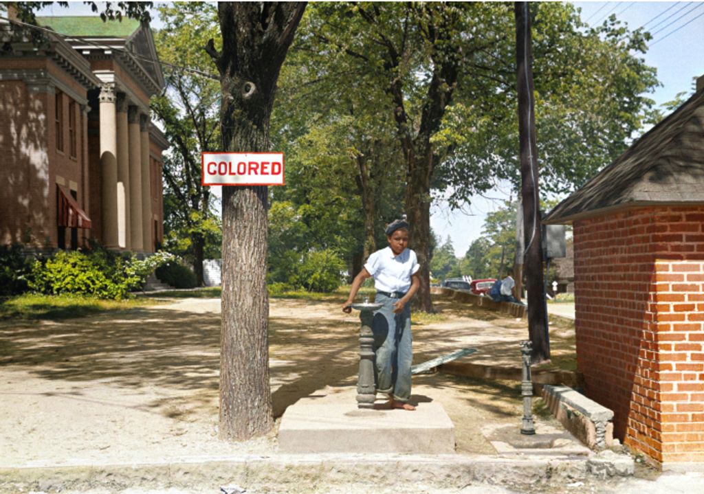 Young black boy drinking from a fountain on the country courthouse lawn in North Carolina next to a sign that says "Colored"