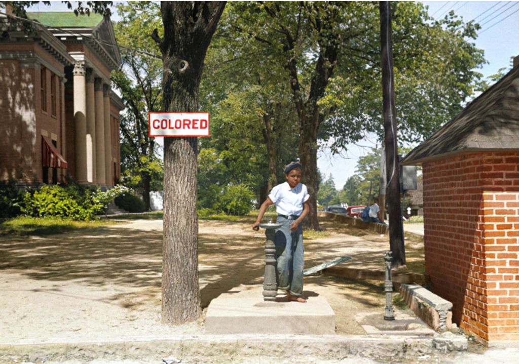 Young black boy drinking from a fountain on the country courthouse lawn in North Carolina next to a sign that says "Colored" 