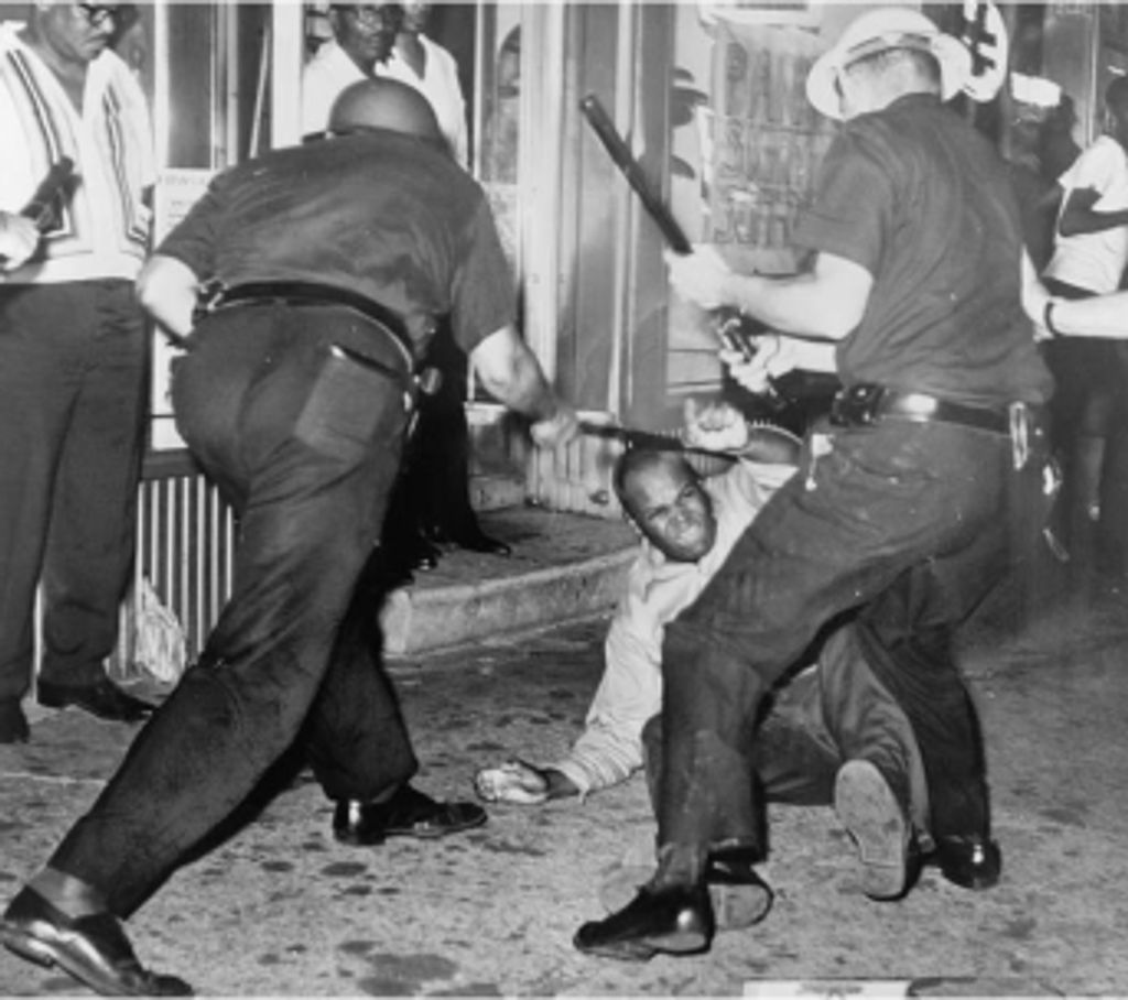 A black and white photograph of two white policemen brutally beating a black man helpless on ground, with batons