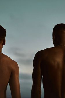 Two shirtless black, young men stand facing stormy beach sky