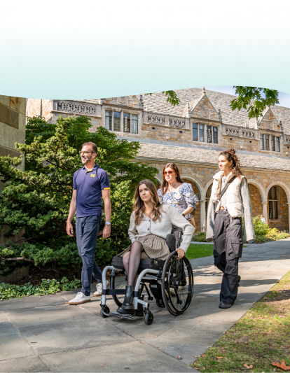 A family of four is walking on a wooden boardwalk through a lush, green forest as sunlight filters through the trees. A young girl in a power wheelchair smiles as she leads the way, accompanied by an adult man and woman, and a young boy who walks beside them holding a walking stick.