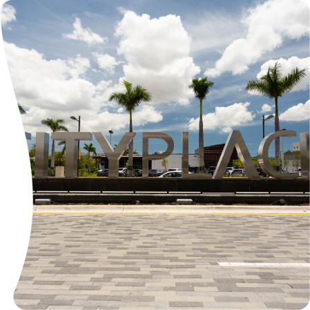 A large metal sign spelling out "CITYPLACE" with palm trees and a modern building in the background. The sky is blue with white clouds.