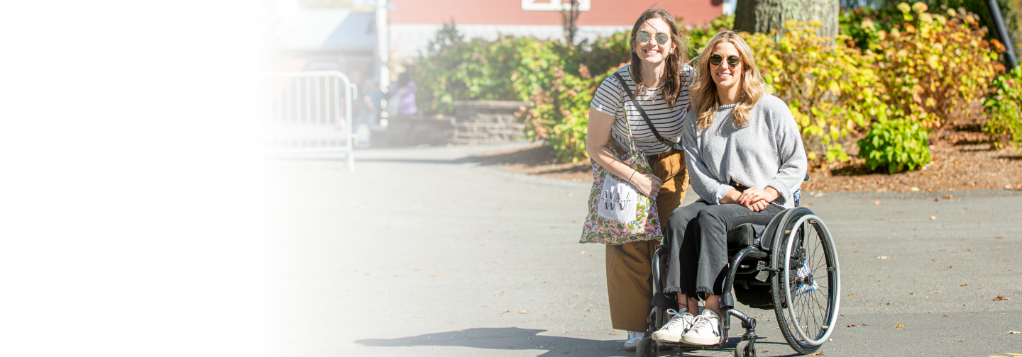 Two smiling women pose outdoors in front of a red barn, one standing and the other seated in a wheelchair, surrounded by greenery and autumn leaves.