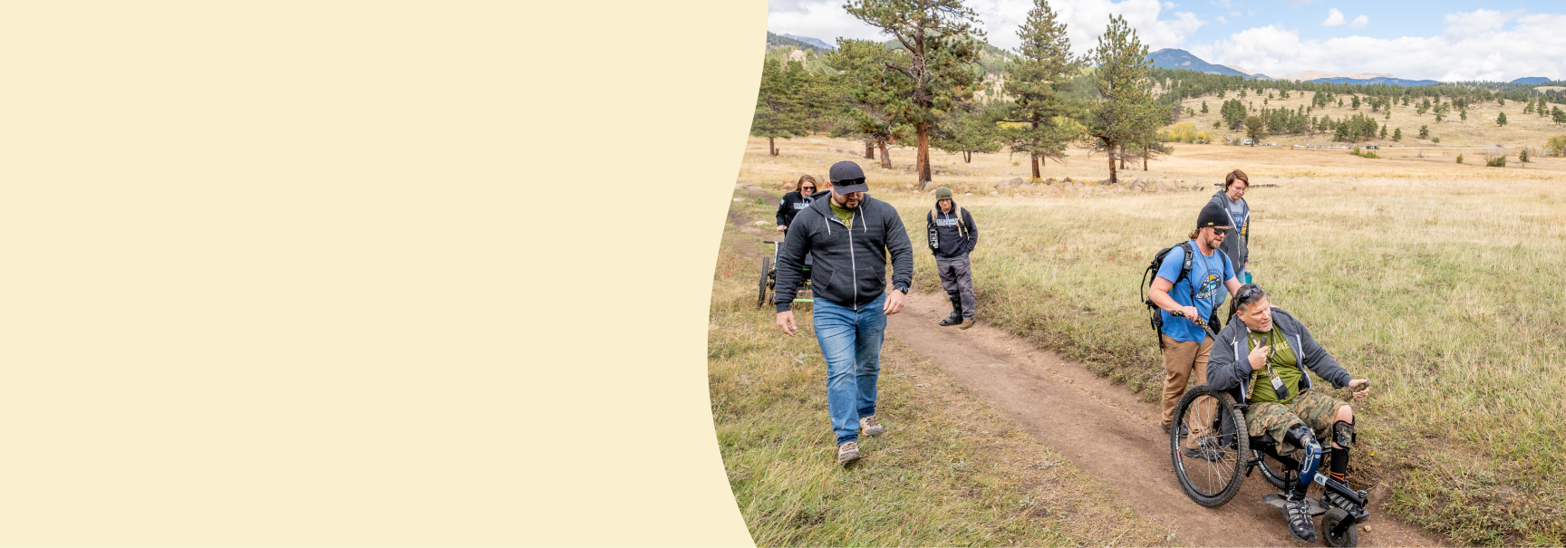 A group of hikers, including a man using an adaptive wheelchair, traverses a scenic trail in Estes Park, Colorado. The path winds through a golden grassy field surrounded by pine trees and rolling mountains under a partly cloudy sky.