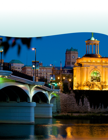 Nighttime cityscape featuring a beautifully lit bridge and an illuminated historic building with a statue on top, surrounded by urban architecture.