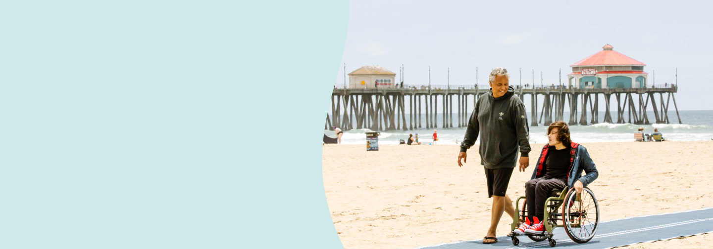 A man walks alongside a child in a wheelchair on an accessible beach path at Huntington Beach, California. The sandy beach and iconic pier with colorful rooftops are visible in the background, with the ocean waves rolling in under a bright sky.