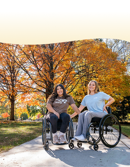 Stef and Erika smile as they sit in their wheelchairs on a paved path in Grand Rapids, each resting a hand on the other’s wheelchair. Vibrant autumn trees with golden leaves frame the scene, creating a warm and inviting atmosphere.