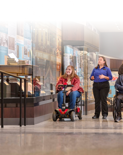 Four people exploring a museum exhibit, including a guide and two individuals using a mobility scooter and a wheelchair, along accessible pathways with informative displays.