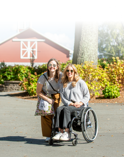 Two smiling women pose outdoors in front of a red barn, one standing and the other seated in a wheelchair, surrounded by greenery and autumn leaves.