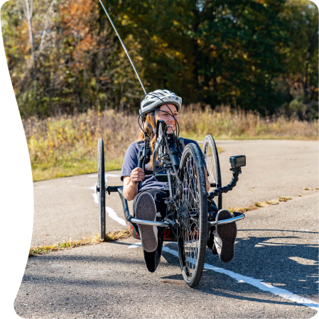 A person wearing a helmet rides a handcycle along a paved path, surrounded by autumn foliage. They propel forward, enjoying the outdoor setting.