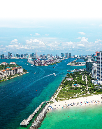 A panoramic view of Haulover Beach Park in Miami, Florida. The image shows a large, green park area with palm trees. The ocean is in the background with blue water and whitecaps.