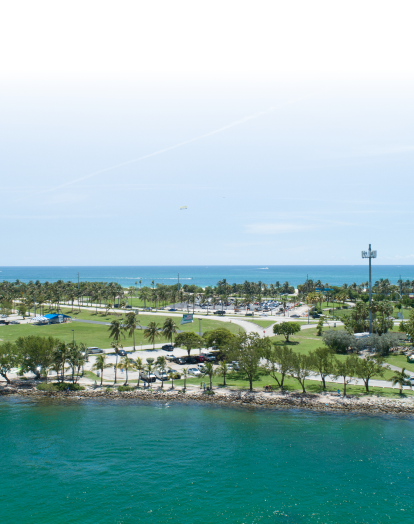 A panoramic view of Haulover Beach Park in Miami, Florida. The image shows a large, green park area with palm trees. The ocean is in the background with blue water and whitecaps.