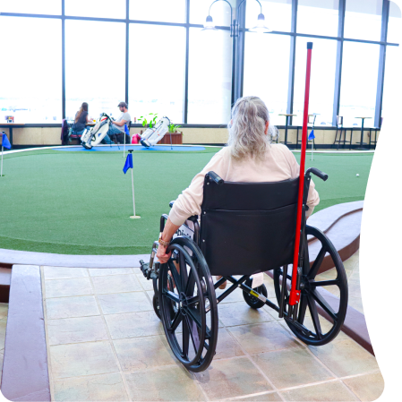 A person in a wheelchair approaches an indoor putting green at Palm Beach Airport, with other travelers visible in the background.