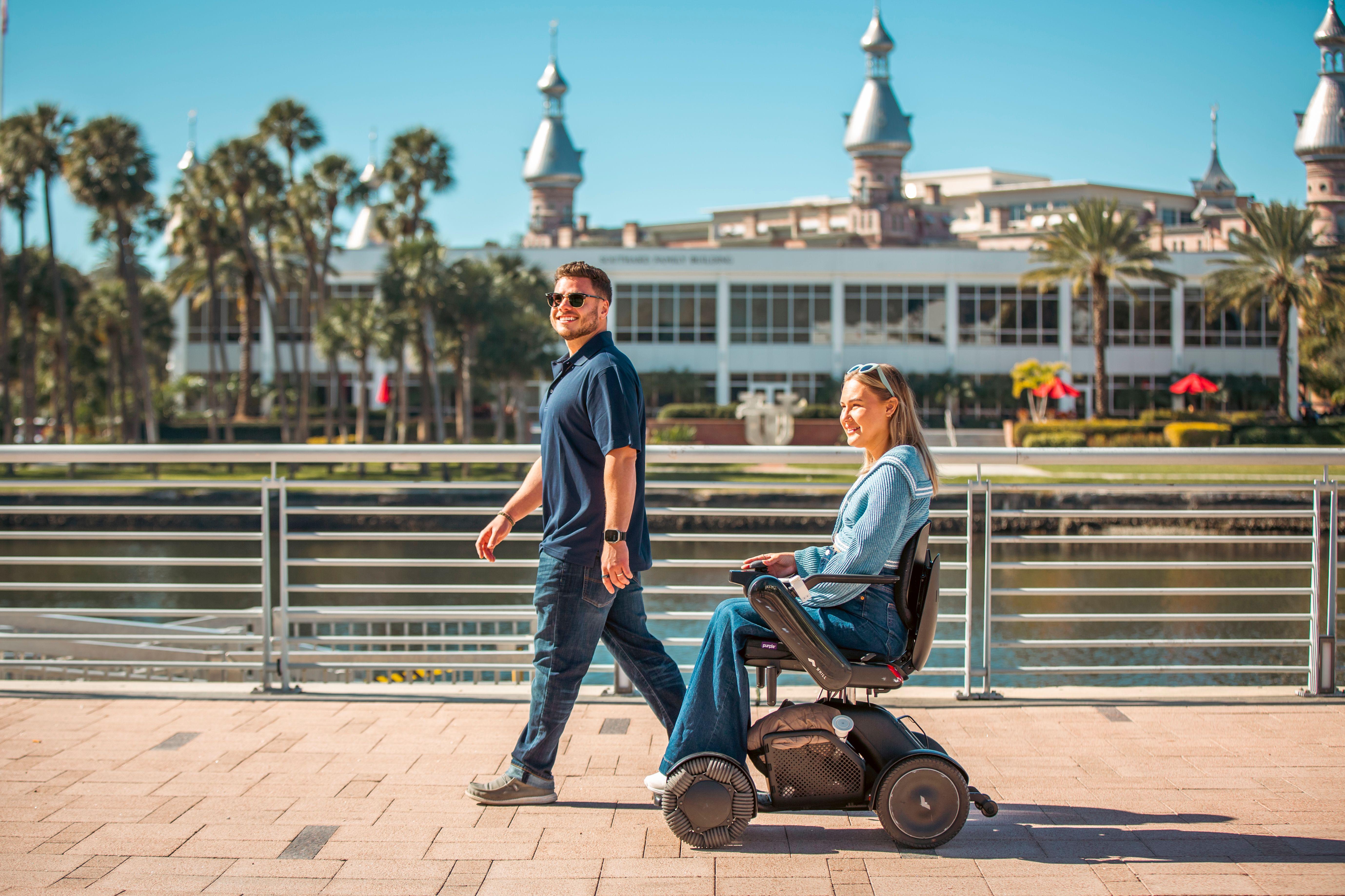 A woman using a WHILL power wheelchair moves along a sunny waterfront promenade beside a man who is walking, with palm trees and silver minarets in the background.