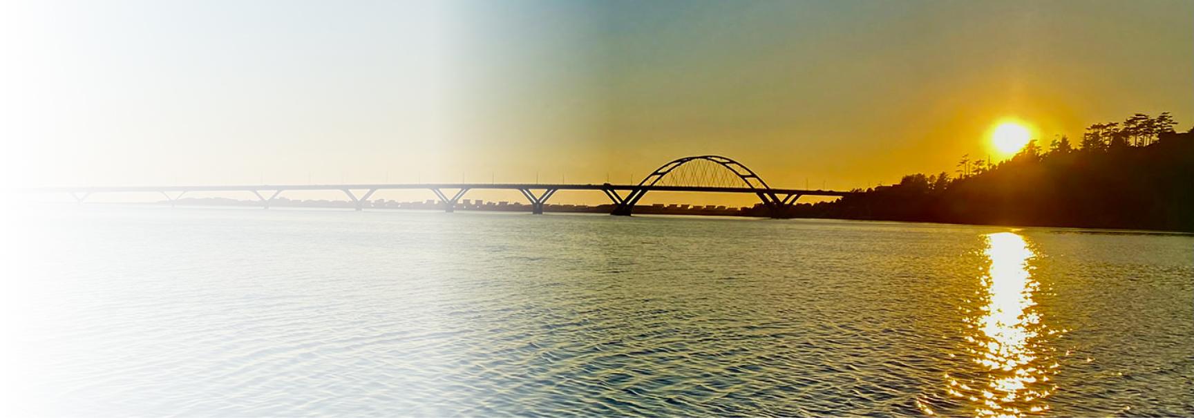 A serene sunset over Alsea Bay in Waldport, Oregon, with the Alsea Bay Bridge silhouetted against the golden sky. The sun reflects off the calm water, creating a warm and tranquil scene with a sandy shore in the foreground.