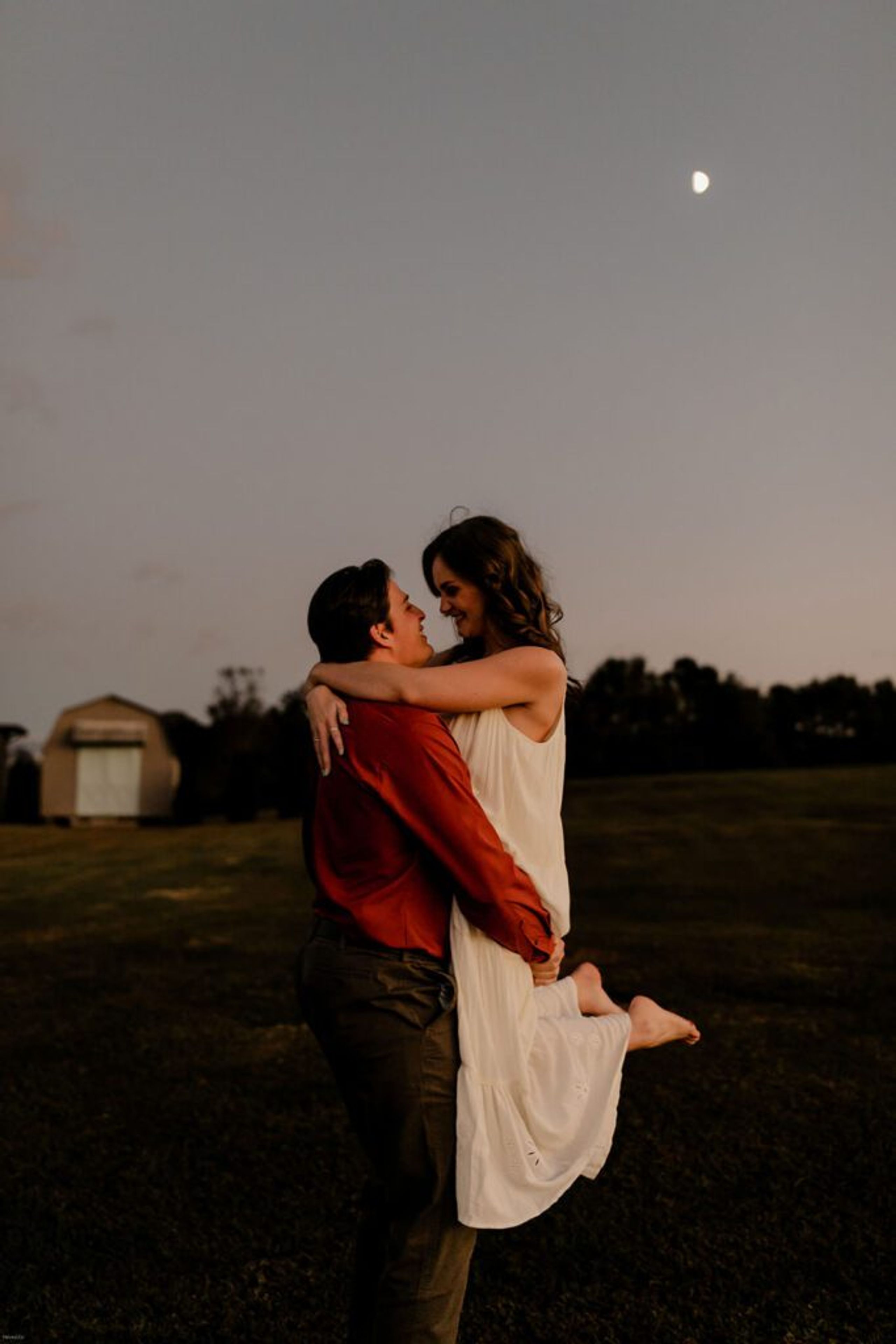 Couple embracing at dusk during their engagement photoshoot, with the moon rising in the background.