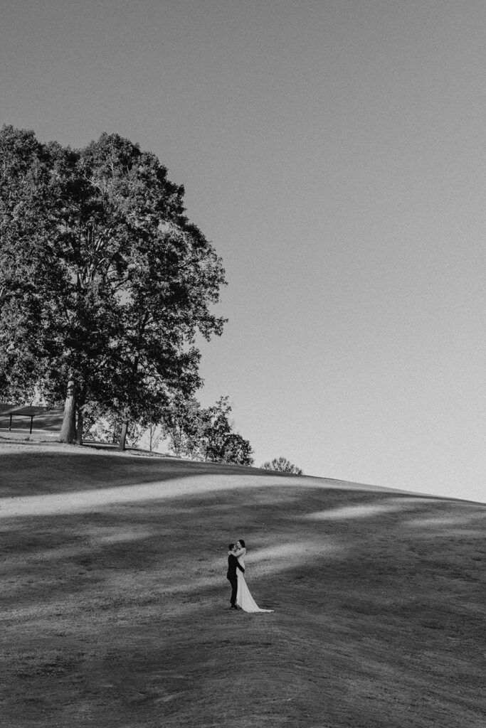 Black and white photo of a bride and groom embracing on a hill at Chimney Oaks Golf Club under a large tree