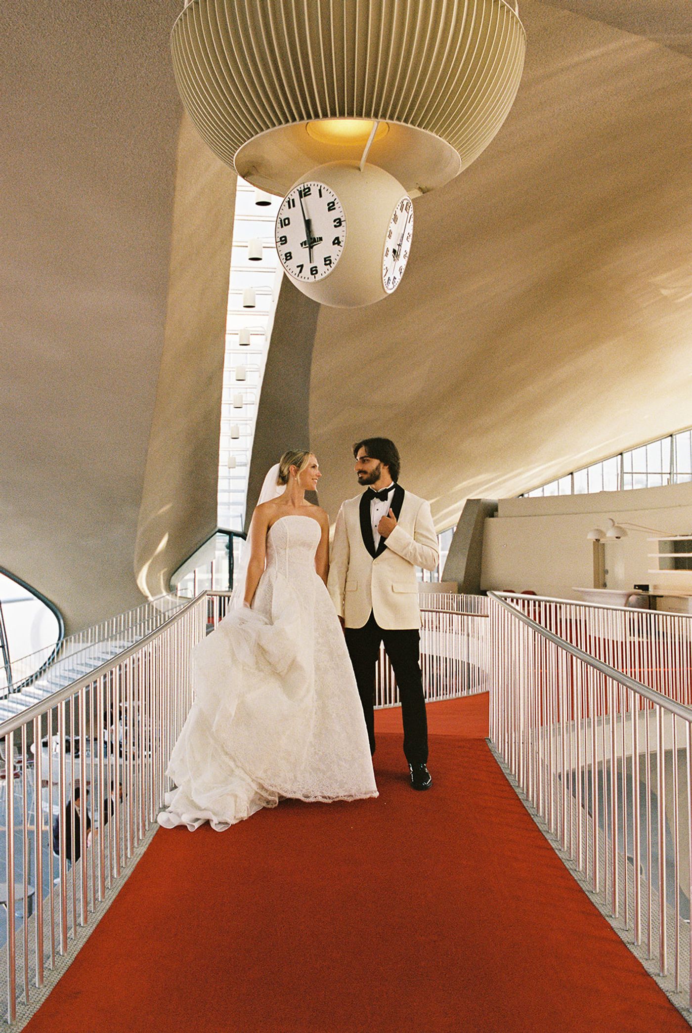 Bride and groom walking the red-carpeted tube under the TWA Flight Center clock
