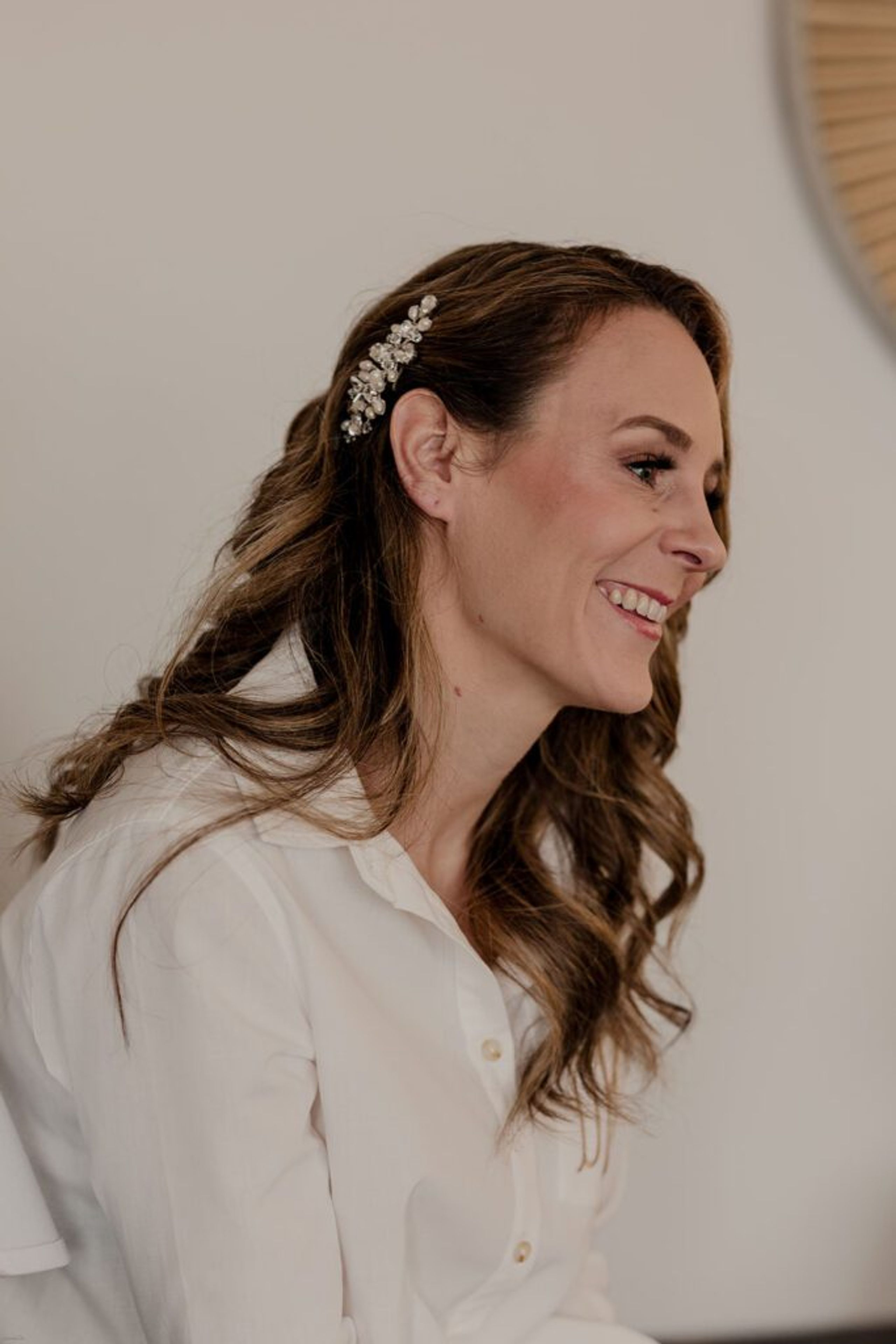 Deb smiles warmly as she prepares for her wedding day, wearing a white shirt with her hair adorned with a delicate hair clip.