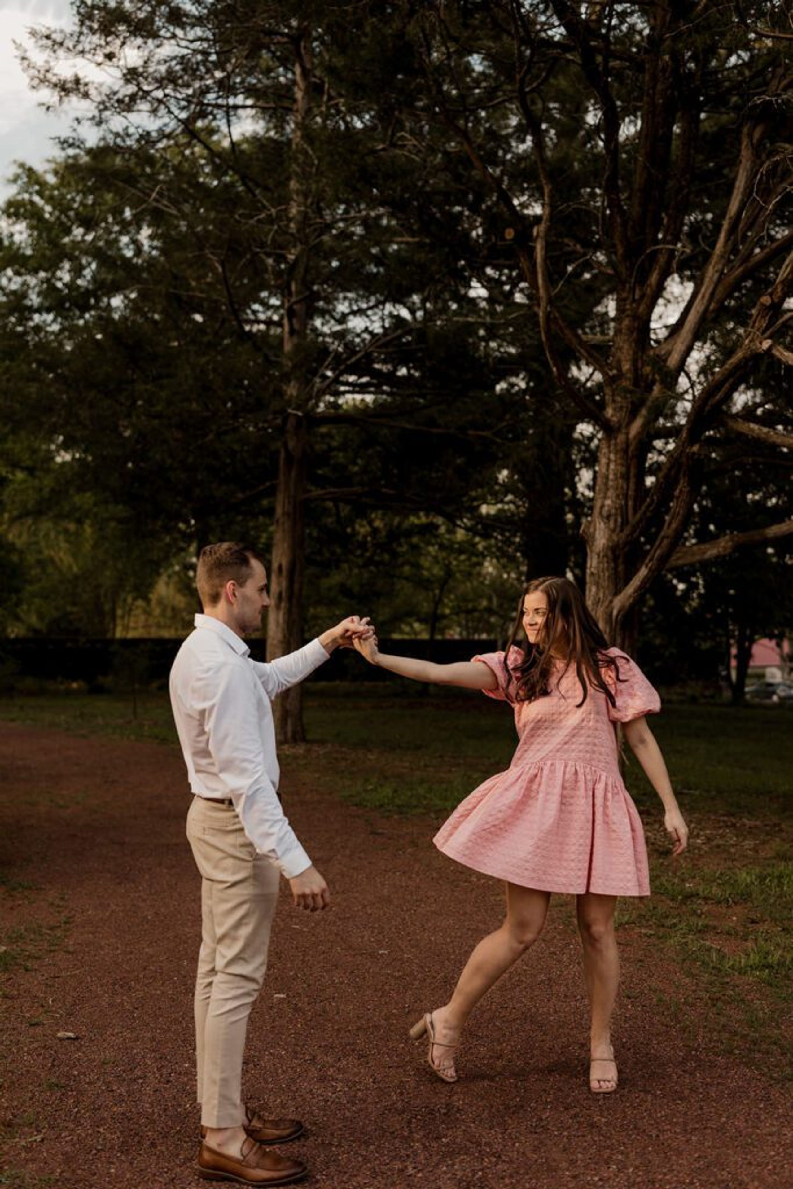 Couple dancing together in a park during their engagement photoshoot, with the woman wearing a pink dress and the man in a white shirt.