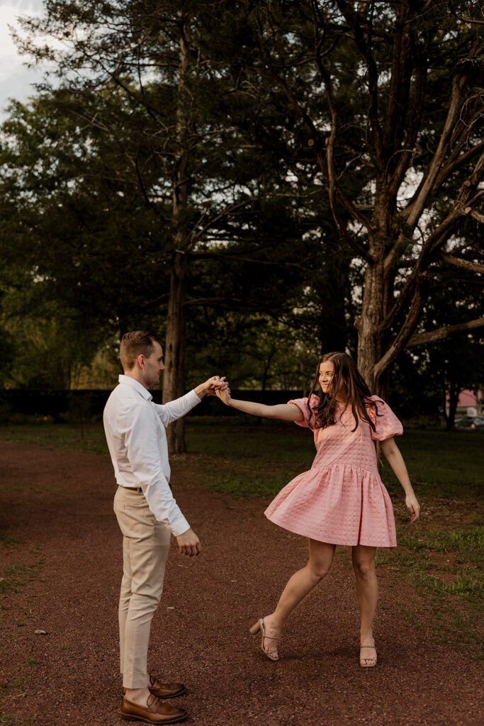 Couple dancing together in a park during their engagement photoshoot, with the woman wearing a pink dress and the man in a white shirt.