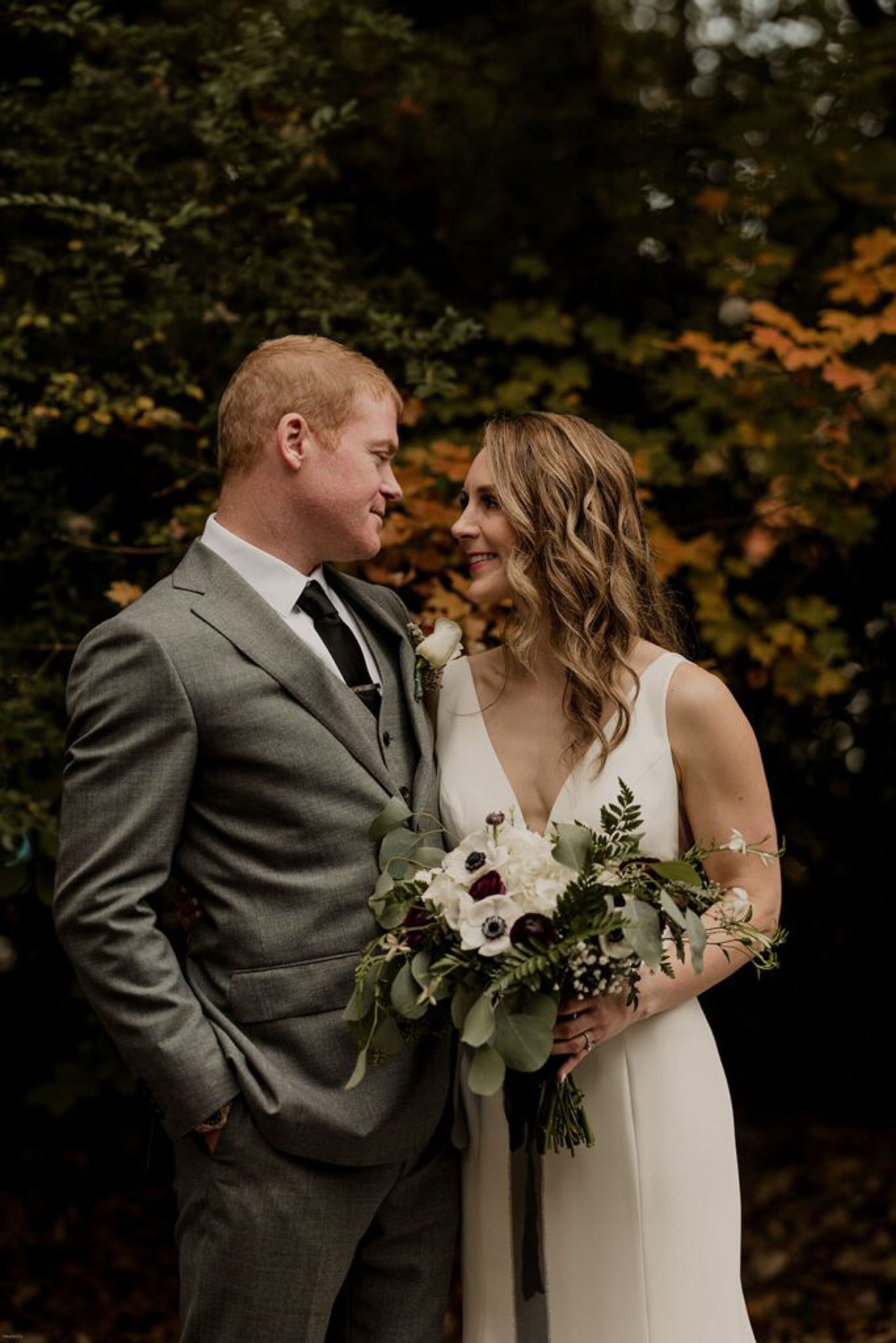 Scott and Deb share a loving gaze, standing close together at 173 Carlyle House with a lush green backdrop, holding Deb’s bouquet of white and burgundy flowers.
