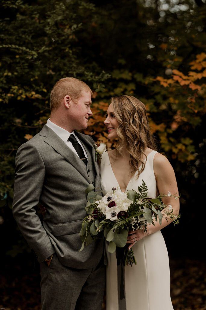 Scott and Deb share a loving gaze, standing close together at 173 Carlyle House with a lush green backdrop, holding Deb’s bouquet of white and burgundy flowers.