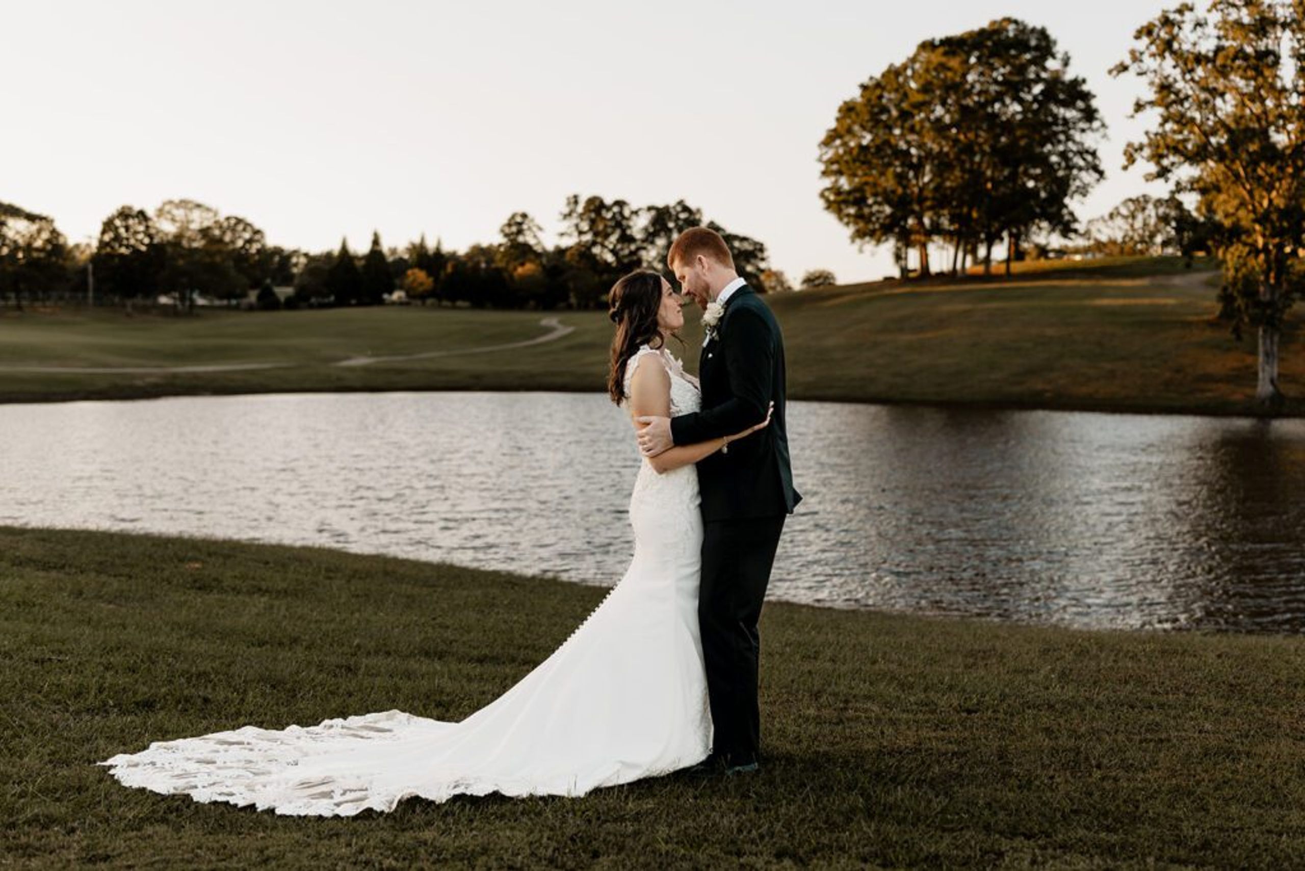 Bride and groom embracing by the lake during a sunset wedding at Chimney Oaks Golf Club, with lush greenery and trees in the background.