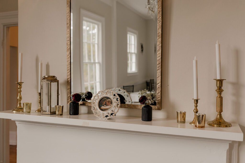 A mantel at 173 Carlyle House decorated with elegant gold candlesticks, a framed photo of Scott and Deb, and small floral arrangements, with a large mirror reflecting the room's bright windows