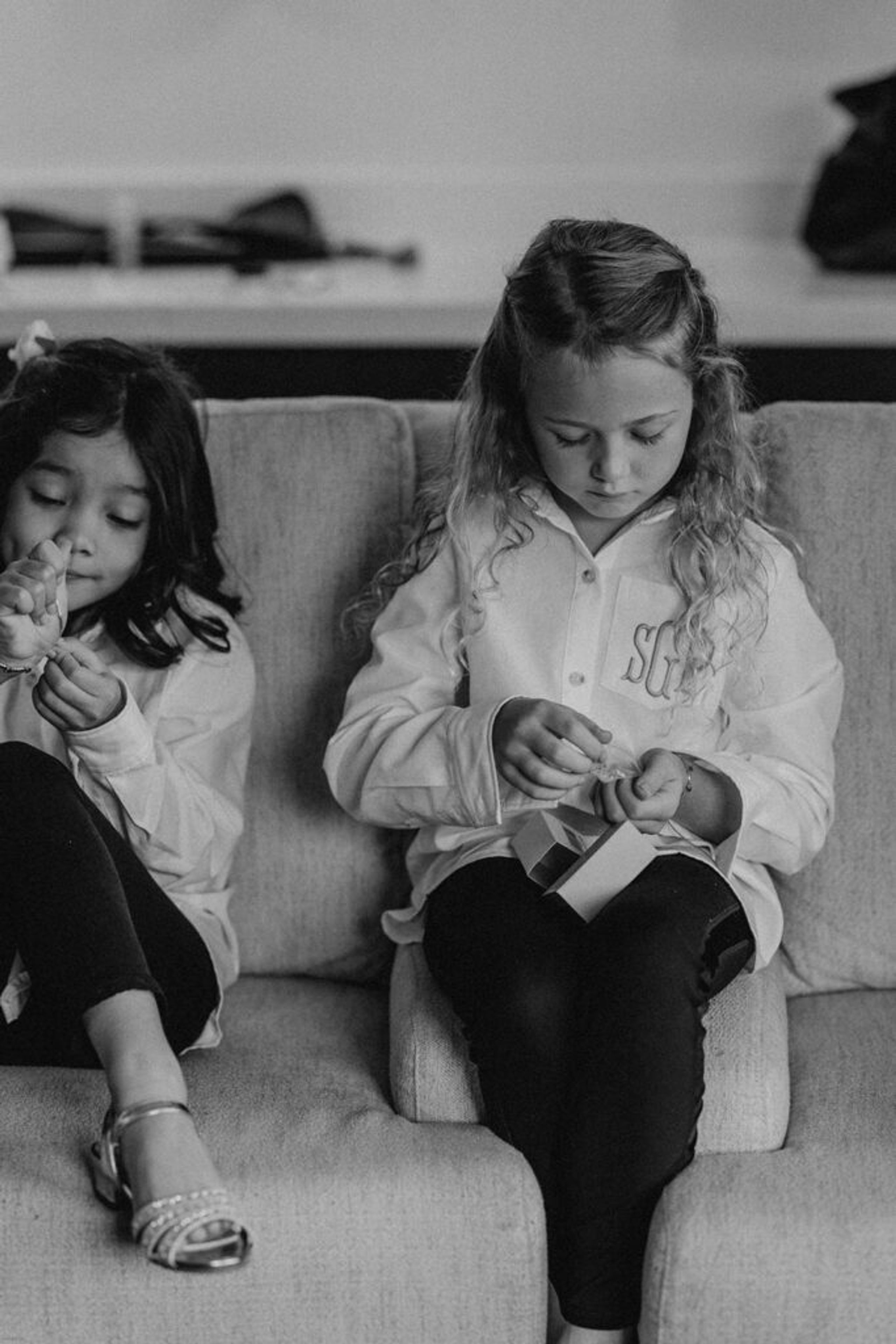 Two young flower girls sit on a couch, focused on opening small gifts before the wedding ceremony at 173 Carlyle House