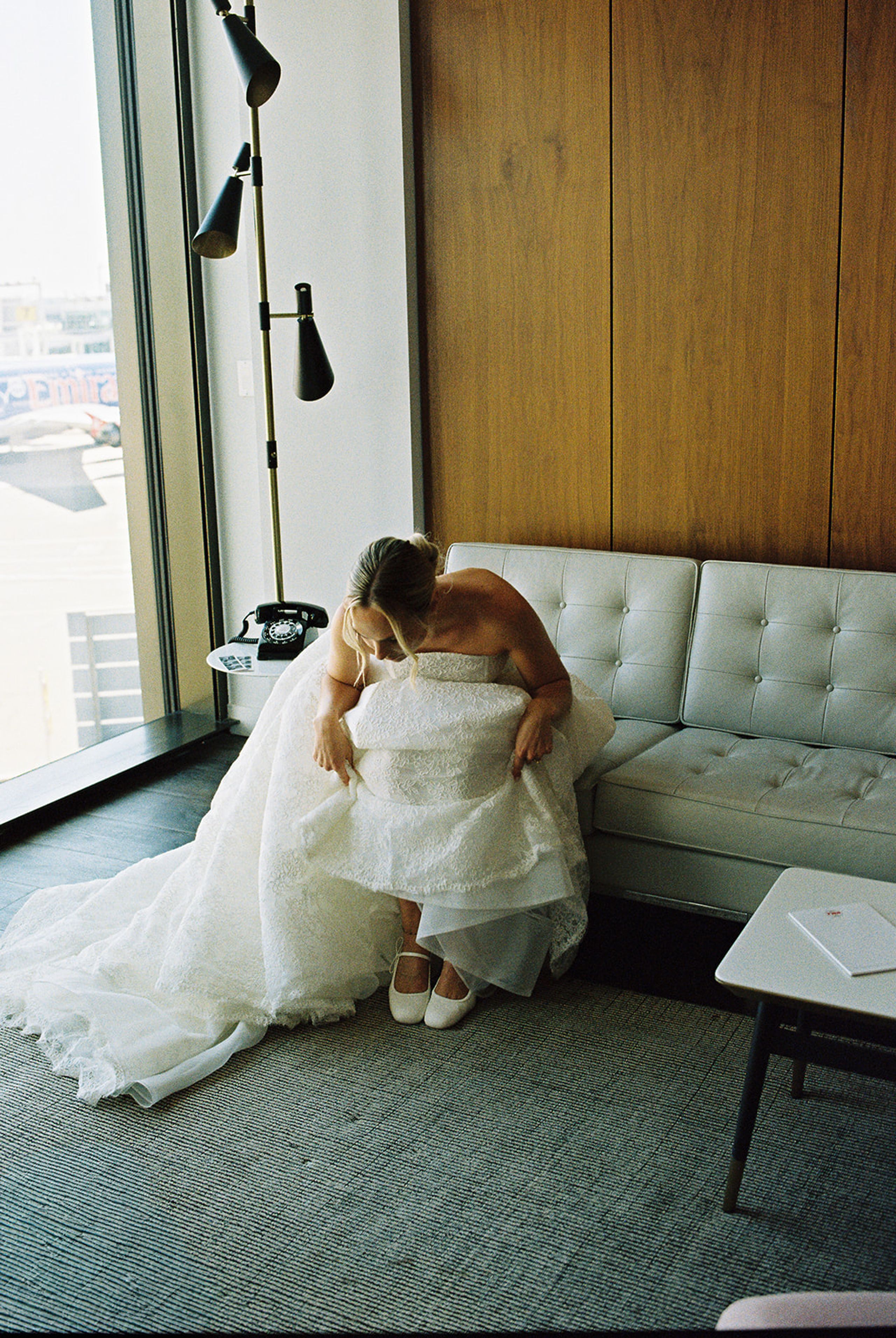 Bride stepping into her lace wedding gown by the window of a TWA Hotel guest room in New York