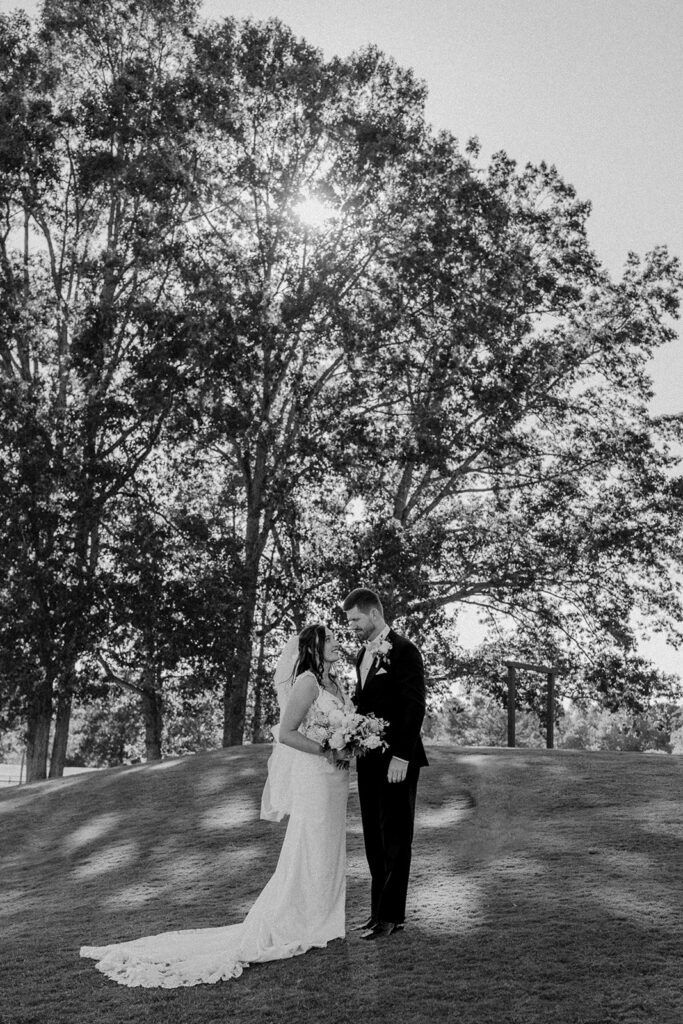 Black and white photo of a bride and groom embracing under a large tree with sunlight filtering through the branches at Chimney Oaks Golf Club