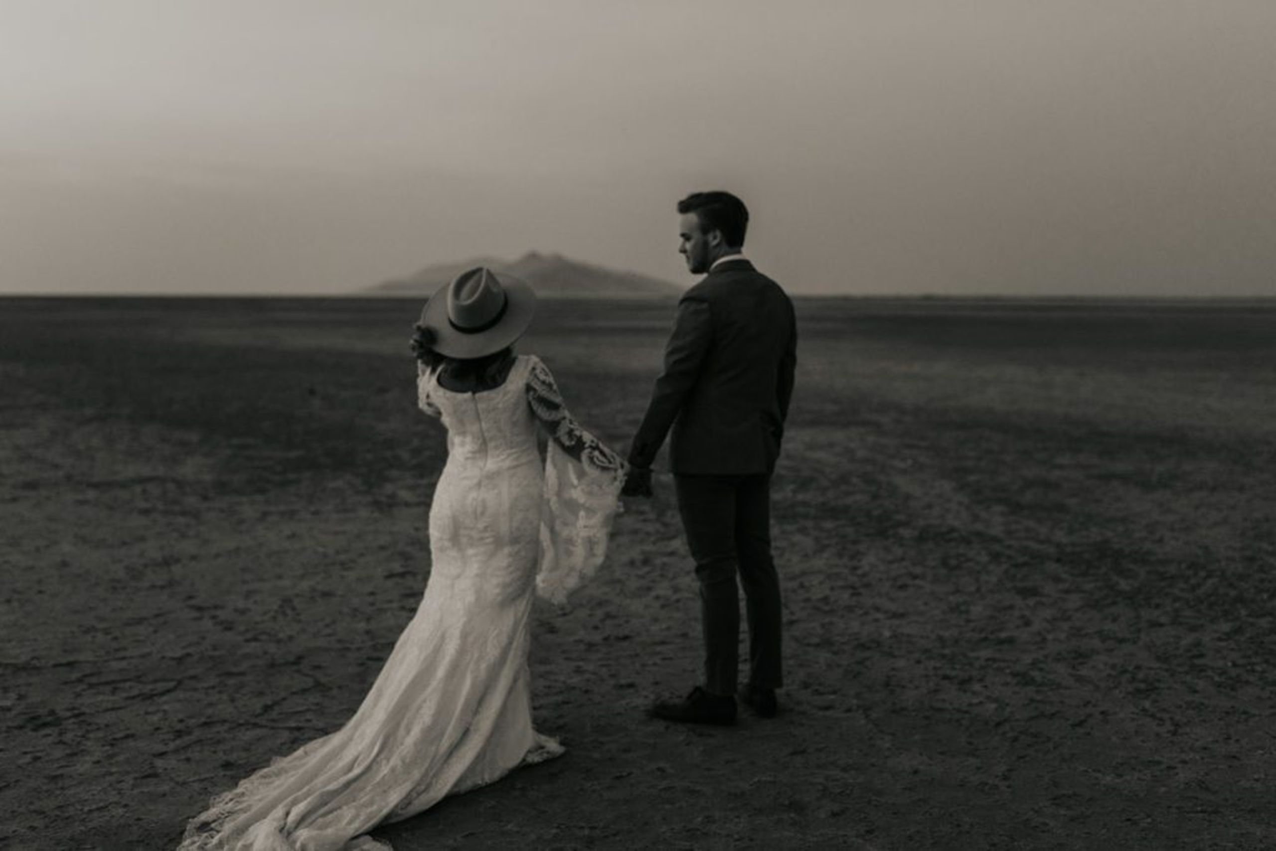 A bride in a lace dress and wide-brimmed hat walks hand in hand with the groom across a vast, open desert landscape, reflecting the adventurous and unique spirit of their Airbnb elopement.