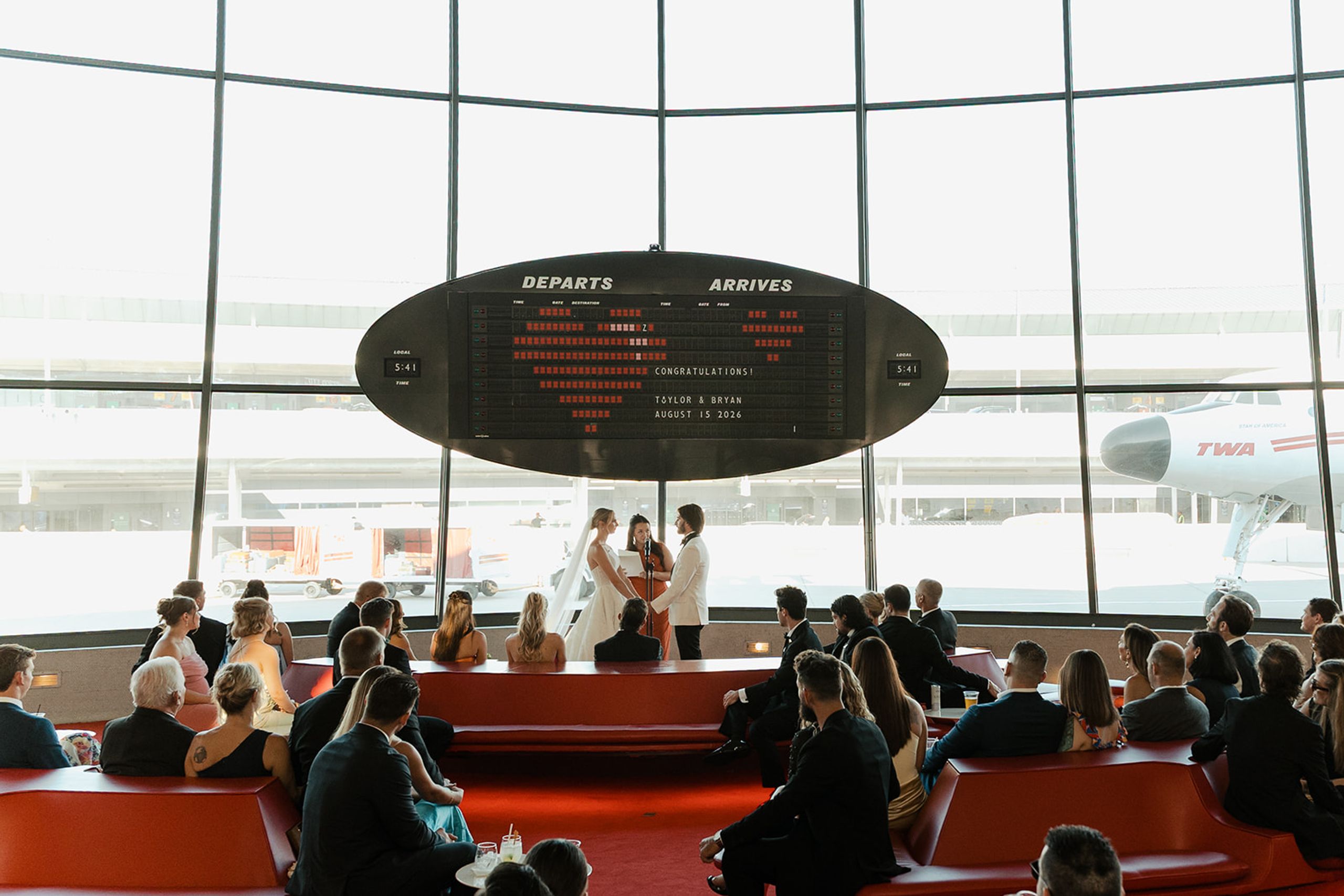 Wide view of a wedding ceremony in the TWA Flight Center with guests seated below the departures board